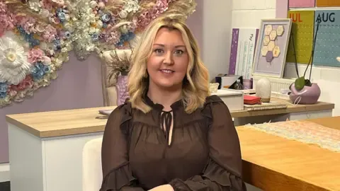 Rebecca is sitting on a chair. She is wearing a black top and has medium blonde hair. To her right is a table. There is a desk behind her. The walls are decorated in colour.
