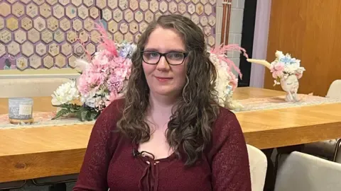 Victoria is sitting. Behind her is a wooden table. She is wearing a burgundy top and has long brown hair. She is wearing black glasses. The wall of tiles is behind her. There are flowers on the table.