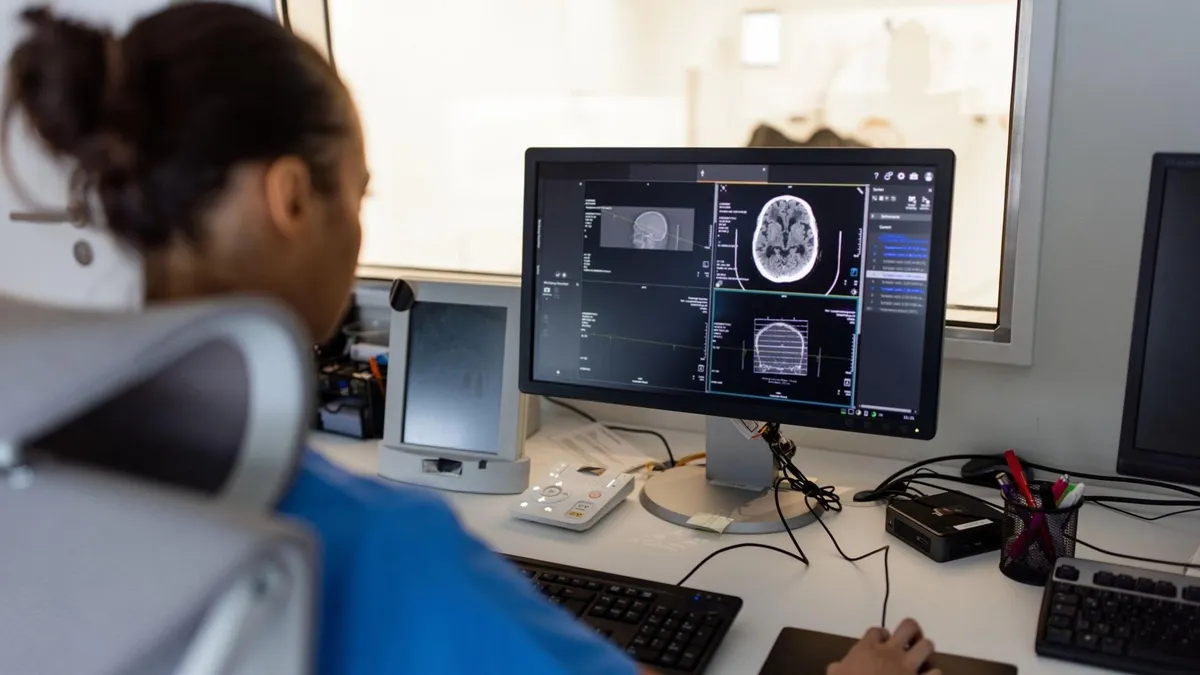 A nurse looks at a brain scan on a computer. In the background, an MRI scanner is visible through a glass window.