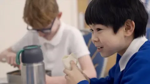 Primary school boy sits in his school canteen in his school uniform smiling whilst eating a wrap.