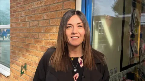 Headteacher of Warter Primary School in York, Helen Houghton smiles into the camera. She's stood outside against a brick wall and is smiling with brunette long hair.