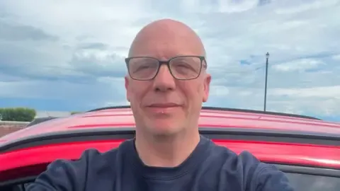 A selfie of a man wearing a navy jumper and glasses. He is standing outside, seemingly in front of a red car.