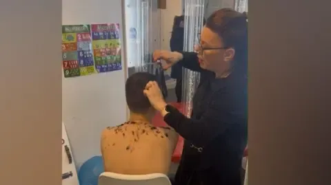 A photo taken from behind of a young boy sitting in a white chair while a woman cuts his hair.