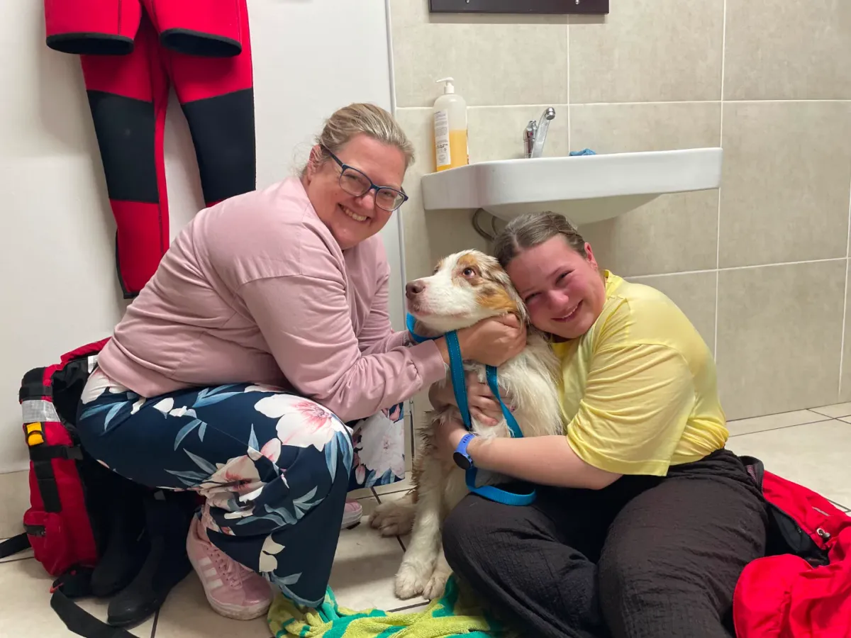 Two women squat in a bathroom holding a small brown and white dog on a leash. Both women are smiling at the camera.