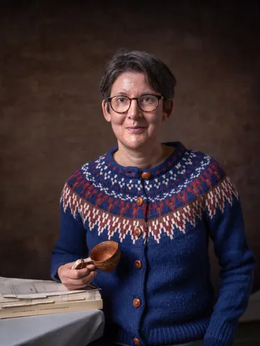 A woman holds a carved wooden cup and rests her hand on a book