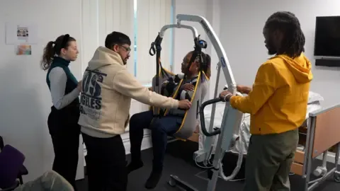 Four new staff are engaged in hoist training. Two of the staff are standing to the left of the mobile hoist and one of the carers, who is wearing a yellow hoodie, is standing to the right and operating the controls. A female staff member is sitting in the hoist. She has her arms crossed across her chest and a male member of staff is supporting the hoist sling in front of her.