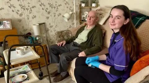Cole-Wilkin, now working as a care assistant, sits inside the home of Stephen Mears. Stephen is sitting in a room with leaf-patterned wallpaper. There is a metal framed walker to the left of the picture with an empty plate and cup on a tray. Mollie is wearing a purple carers uniform with white piping and has blue gloves on.