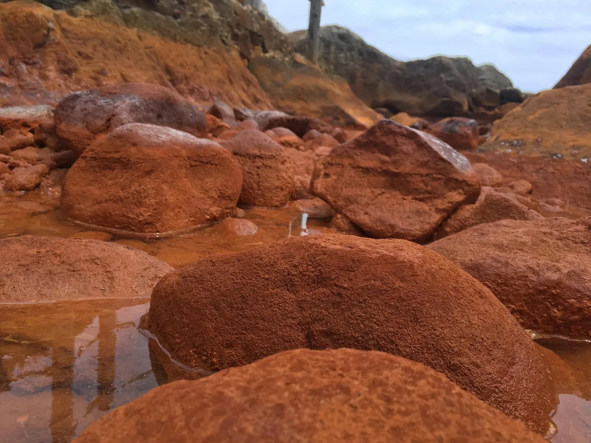 A close-up picture of the sediment and rocks of one of five hot springs during low tide, showing iron oxide mineral precipitates