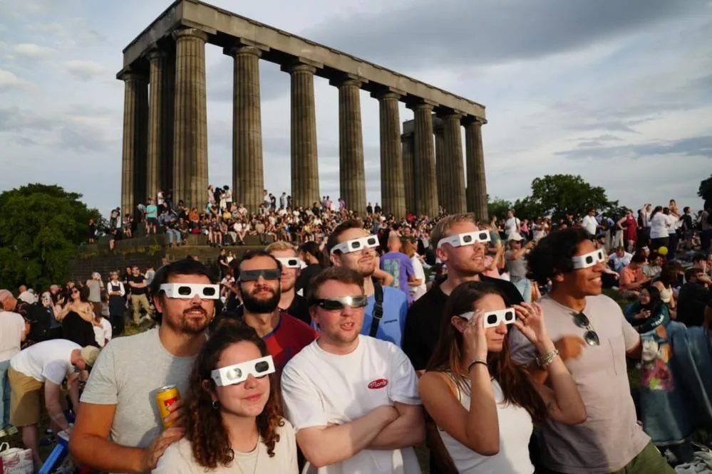 Crowds of mixed genders and ages stand on a grassy hilltop with the columns of the National Monument visible in the background. A group of friends at the front can be seen wearing eclipse glasses.