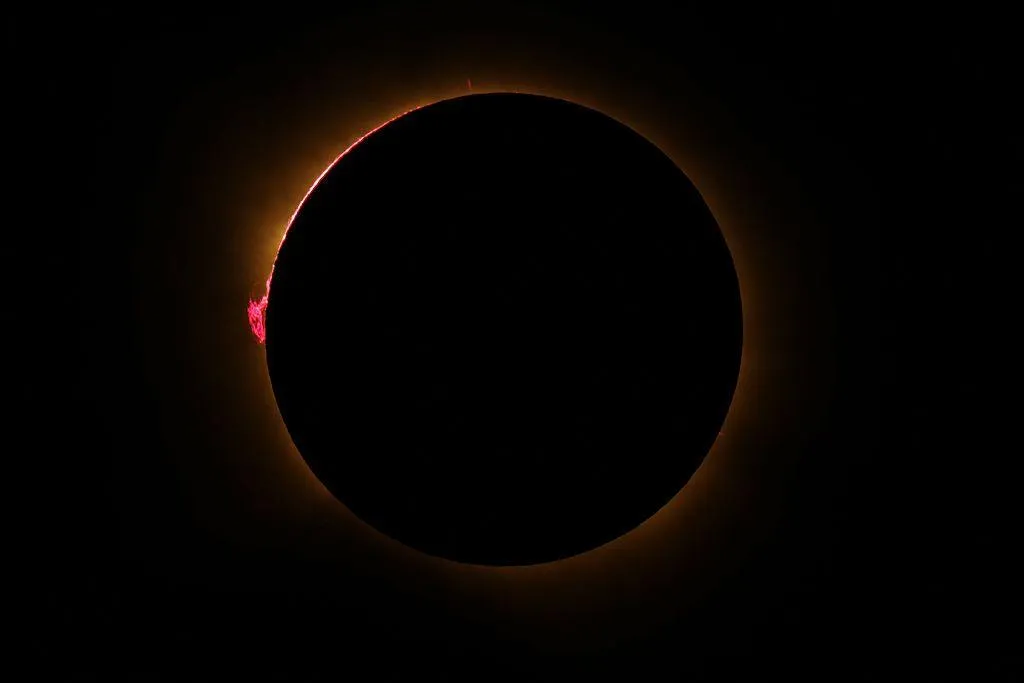 A black darkened sky with a circle of orange light bordering a black circle – the blocked Sun - and small loops of pink can be seen coming from the surface of the Sun
