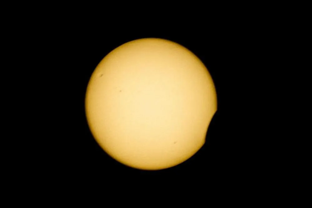 A yellow circle – the Sun – sits against a black sky. There is a small crescent “bite” of black removed from the lower right corner as the Moon moves across it