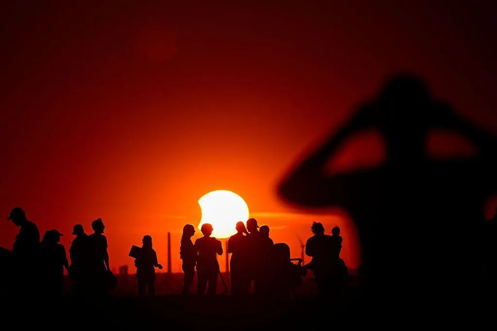 An orange sky with the Sun in the centre, there is a small bite sized chunk taken out of its left side as the Moon covers the surface. People standing around in groups with telescopes are in shadow