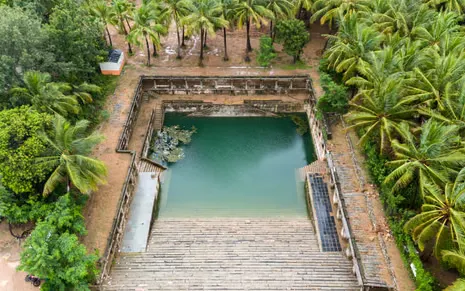 A rectangular pool with steps running down to a square of blue water surrounded by a colonnade and palm trees