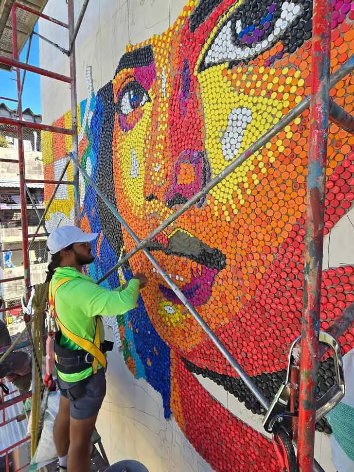 An artist stands in a lift on the side of a building, installing a mural with individual bottle caps