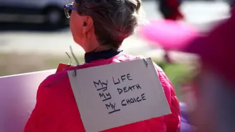 Getty Images A person stands with their back to the camera and a sign hanging over her neck reading 'my life, my death, my choice', outside parliament in London in September.
