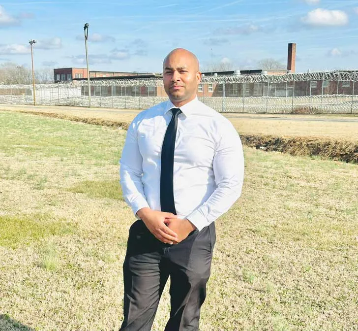 Kerwin Pittman, a Black man in a white button down shirt and black tie, stands in front of a correctional facility.