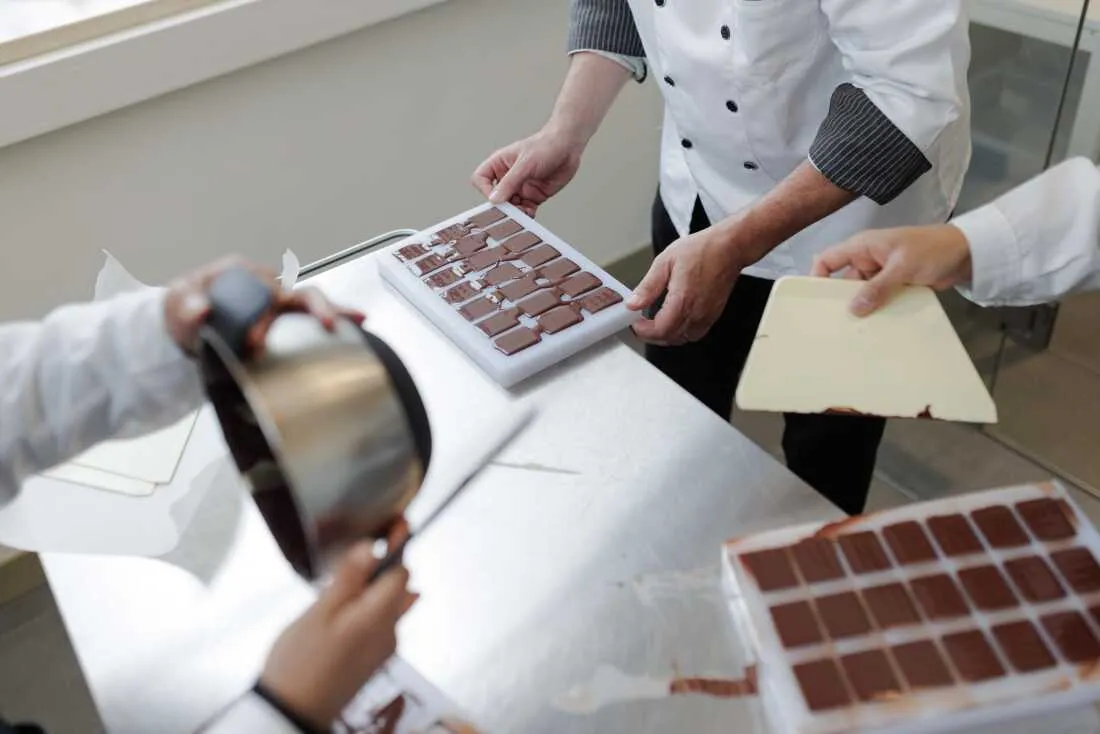 A chef handles molds filled with chocolate as workers prepare artisan bars during a production session in Perugia, May 6, 2026. This final stage of molding requires the trays to be vibrated or tapped to remove air bubbles before being placed in a cooling environment to solidify. The process ensures that the finished chocolate bars have a smooth texture and a uniform appearance