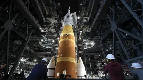 Artemis II sits in the Vehicle Assembly Building at NASA’s Kennedy Space Center as three workers wearing white hard hats look up at the rocket