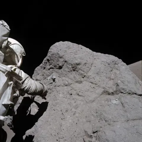 The Apollo astronaut Harrison Schmitt, from Apollo 17, is collecting samples rock on the Moon. The rock is slightly taller than him and about 3 times as wide. He is on the left of the photo with his back to the camera, and is wearing a white NASA space suit as he tries to collect the sample using his right arm. His body is creating a shadow on the large dark grey rock. At the top of the photo you can see the blackness of space.