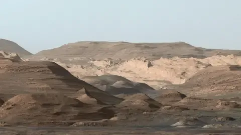 The picture shows the higher regions of Mount Sharp on Mars. In the foreground are some darker areas with some small hillocks with a slightly larger slope off to the left. In the middle of the photo are some lighter more craggy looking rocky areas. In the distance are bigger hills that are more rounded with gentle slopes. The sky, which is visible at the very top is a dark grey.