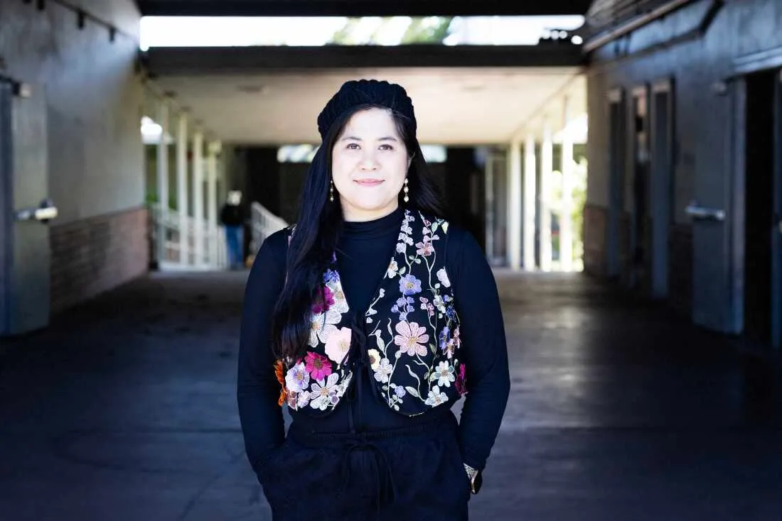 Mary Acebu — a young Filipino woman wearing all black, a flowered vest, and a knitted beret — stands in the middle of an outdoor entryway at Riverview Middle School.