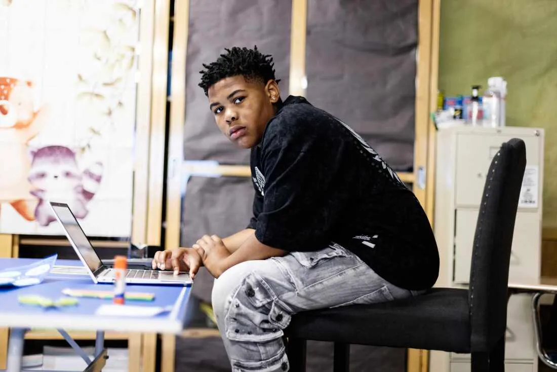 King — a black, eighth grade boy with short locs is wearing a black t-shirt and gray jeans. He looks at the camera while sitting at a laptop working on an assignment in Mary Acebu's class.