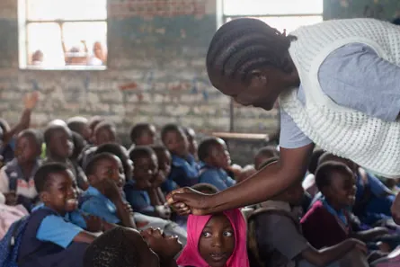 A healthcare worker bends over a seated child to administer a polio drop, while other rows of children look on.