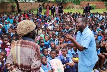 A man in blue healthcare worker scrubs speaks to a woman in front of a large group of children.