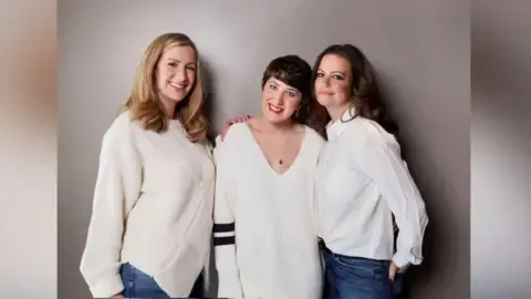Claire Wood The original line up of the the BBC's You, Me and the Big C podcast. Left to right: Rachael Bland, Lauren Mahon and Dame Deborah James. All three are smiling to camera. Rachael is wearing a white jumper and jeans, Lauren is wearing a white jumper with two black stripes on the sleeves and Deborah is wearing a white shirt and jeans.