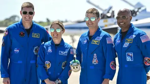 Four Artemis II astronauts stand side by side on a sunny runway, posing for a group portrait. They all wear bright blue NASA flight suits covered in mission patches and name badges, with dark boots. One astronaut in the centre holds a small mascot or model in both hands. Behind them, two sleek white-and-blue T‑38 training jets sit on the concrete, their pointed noses facing left and right, with the NASA “meatball” logo visible on a tail fin. The sky above is clear and pale blue, giving the scene a crisp, formal but upbeat feel.