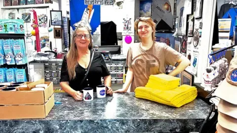 Inside a small, crowded NASA gift shop, two people stand behind a grey marble-effect counter. Shelves and walls around them are packed with space souvenirs, mission posters, and astronaut photos. On the left are boxes of mugs; the woman holds two white mugs decorated with NASA-style logos. Next to her, a younger person in a pale T‑shirt leans on the counter near two neat piles of bright yellow folded T‑shirts. To the right, a rack displays beige and orange NASA baseball caps and small astronaut toys, giving the scene a busy, colourful, fan-filled atmosphere.