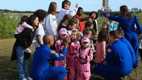 On a grassy patch near water, three Artemis II astronauts in bright blue flight suits crouch down to talk with a group of young children. The children in the centre wear miniature pink spacesuits and caps, facing the astronauts and giving them high‑fives. Other children and parents cluster around them, some holding toddlers, forming a loose semicircle. In the distance, partly blurred, a tall rocket and launch tower rise above the trees. The mood is warm and playful, with astronauts and families smiling and interacting at eye level, turning a serious mission into a friendly, down‑to‑earth moment.