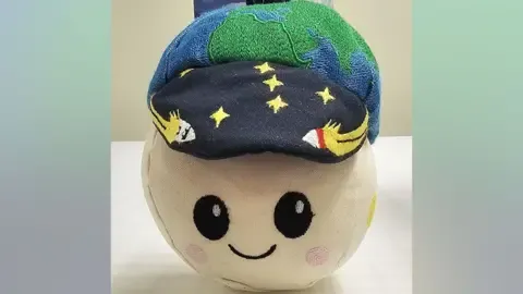A small, round soft toy sits on a white tabletop, about the size of a large grapefruit. Its fabric face is pale cream with big black embroidered eyes, rosy pink cheeks and a simple smiling mouth, giving it a cute cartoon look. On top, it wears a dark blue cap sprinkled with embroidered yellow stars and tiny white rockets. Above the cap is a padded green‑and‑blue globe, like a miniature Earth, with a short dark loop for hanging. The toy’s overall impression is friendly and playful, like a cheerful mascot for a children’s space adventure.
