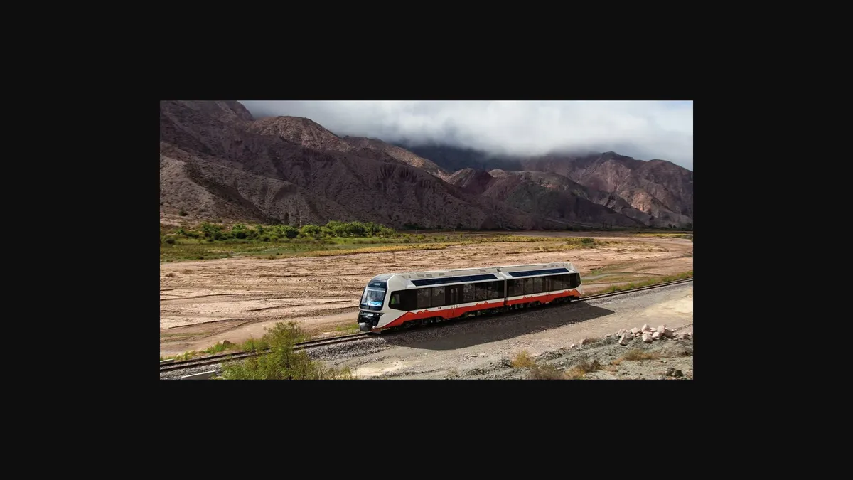 A train with two cars is parked at a station platform, with colorful mountains in the background.