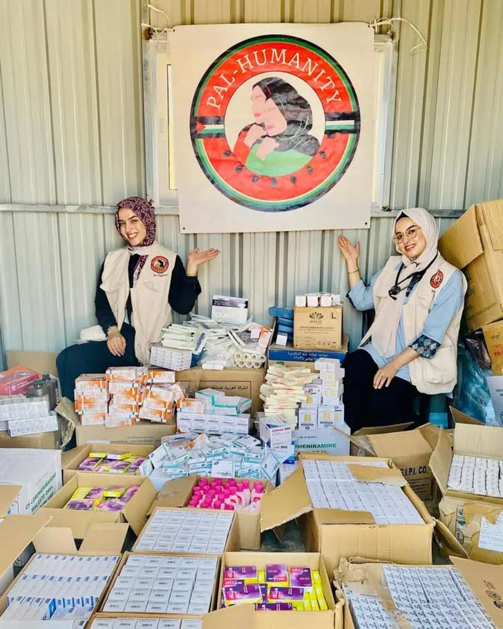 Two young doctors in hijabs smiling in front of boxes of donated medical supplies, under a banner for their nonprofit, Pal Humanity