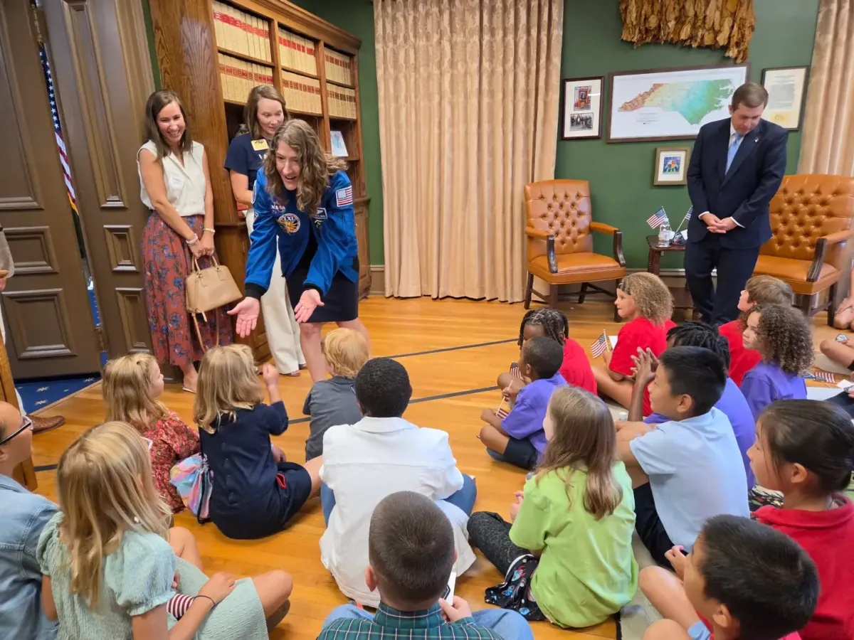 A NASA astronaut in a blue flight suit sits on the floor speaking with a group of children in an ornate green room with wood paneling, bookshelves, and leather chairs. Two women stand nearby observing, while a man in a dark suit stands in the background.
