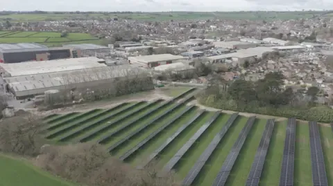 SunGift Solar Aerial view of solar panels on green field near factory and homes.