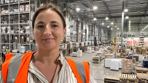 Woman with brown hair wearing a high viz stood in factory with shelves in the background.