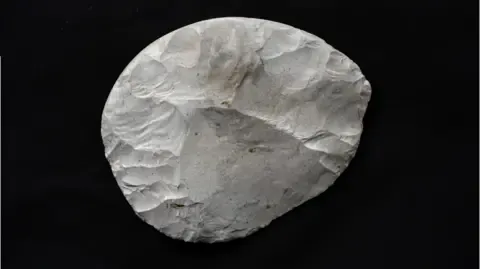 A rounded piece of flint that has been carefully worked to create a knife. It is white, with some brown and grey marks, and is set against a black background. The areas where pieces of the flint have been struck off using another stone are clearly visible. It is not a perfect circle but its sides are rounded.