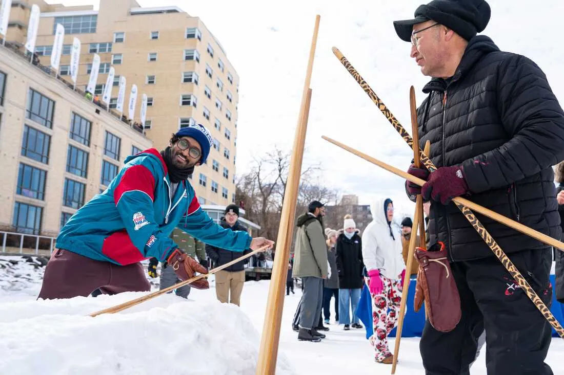 Bill Quackenbush, of Ho-Chunk Nation (right), teaches Praneeth Kolli (left) how to compete in the snow snake competition during the city's annual Frozen Assets Festival on Lake Mendota. Snow snake is a traditional Indigenous winter sport where players slide handcrafted wooden sticks down a trench made of snow for distance.