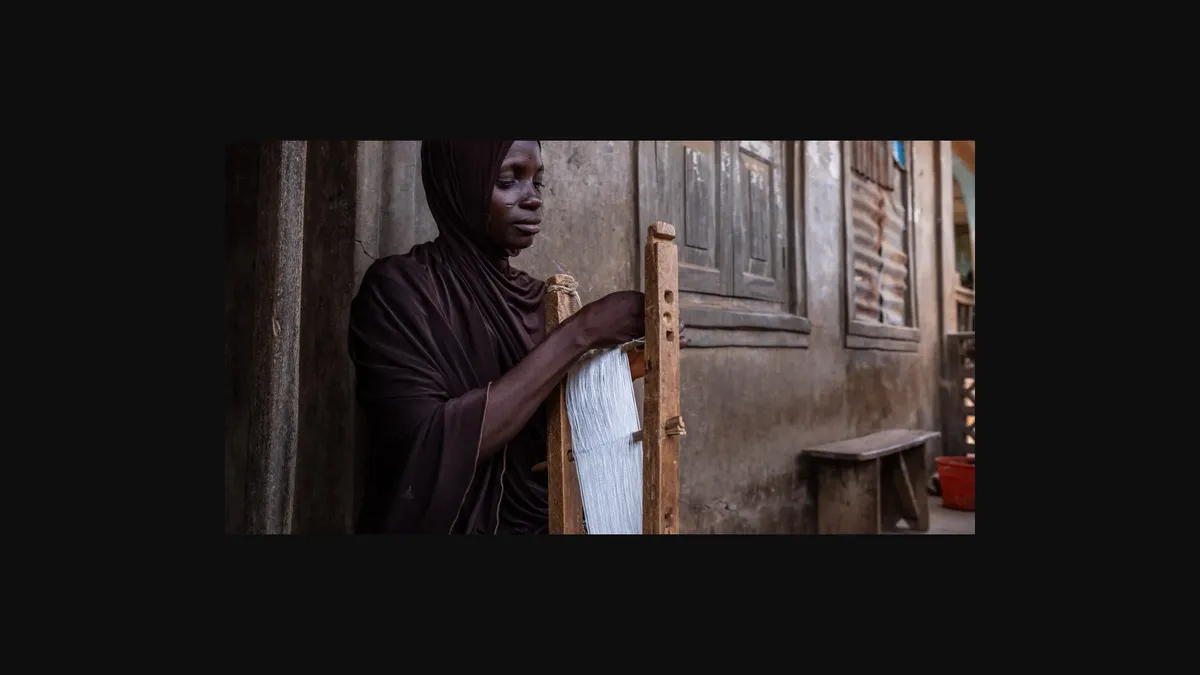 A man sits at a wooden loom, weaving colorful threads.