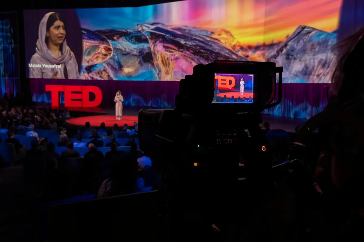 Malala, dressed in pink, stands on stage for TED Talk, standing on red carpet with the TED logo in the background. Viewed from behind a camera in the back of the theater