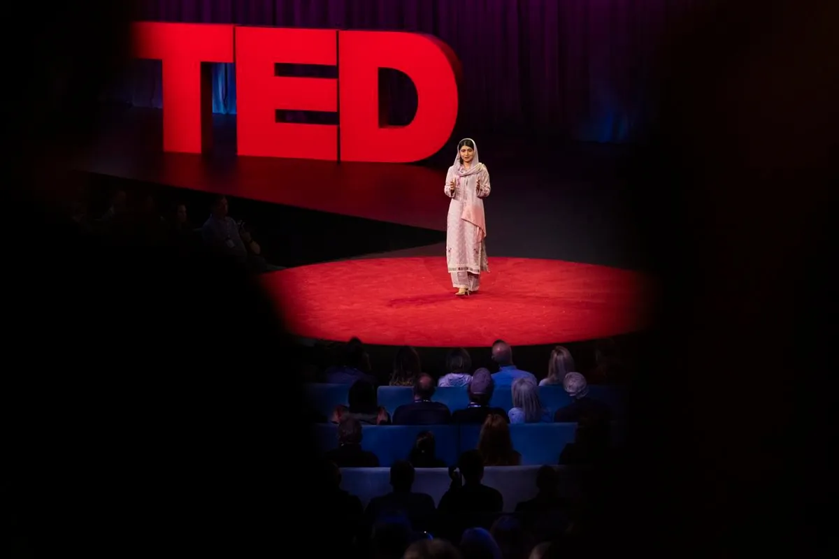 Malala, dressed in pink, stands on stage for TED Talk, standing on red carpet with the TED logo in the background.