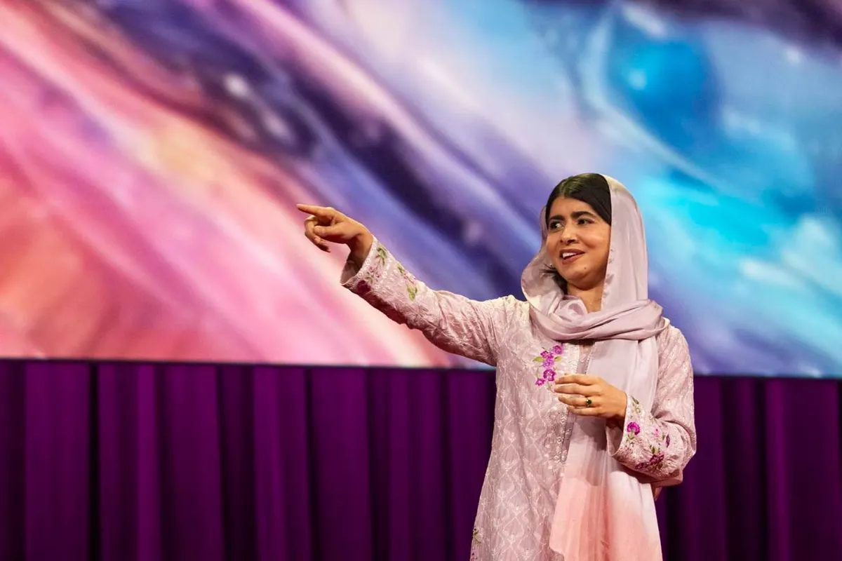 Malala, dressed in pink, stands on stage for TED Talk, points