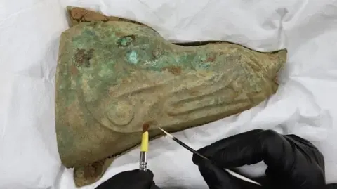 A boar's head standard on its side, with its nose facing to the right. Two black-clad hands can just be seen at the bottom holding a thicker brush on the left and a far finer bush in the right hand.