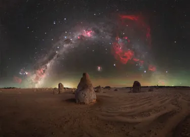A calm, windless night in the Pinnacles desert, as the main limestone pillar aligns almost perfectly with the South Celestial Pole, anchoring the image beneath the rotating sky. To the right, the faint but unmistakable red glow of the Gum Nebula, a vast supernova remnant, adds depth and scale to the scene