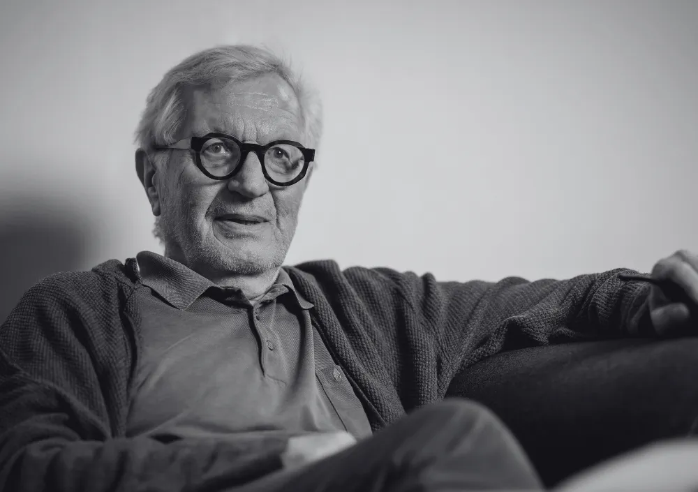 An old castle with a tower and red roof sits on a hill surrounded by trees under a cloudy sky.